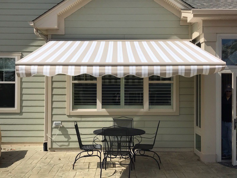 Beige and white striped retractable awning fully extended over a stone patio with black iron dining table and chairs against green house siding