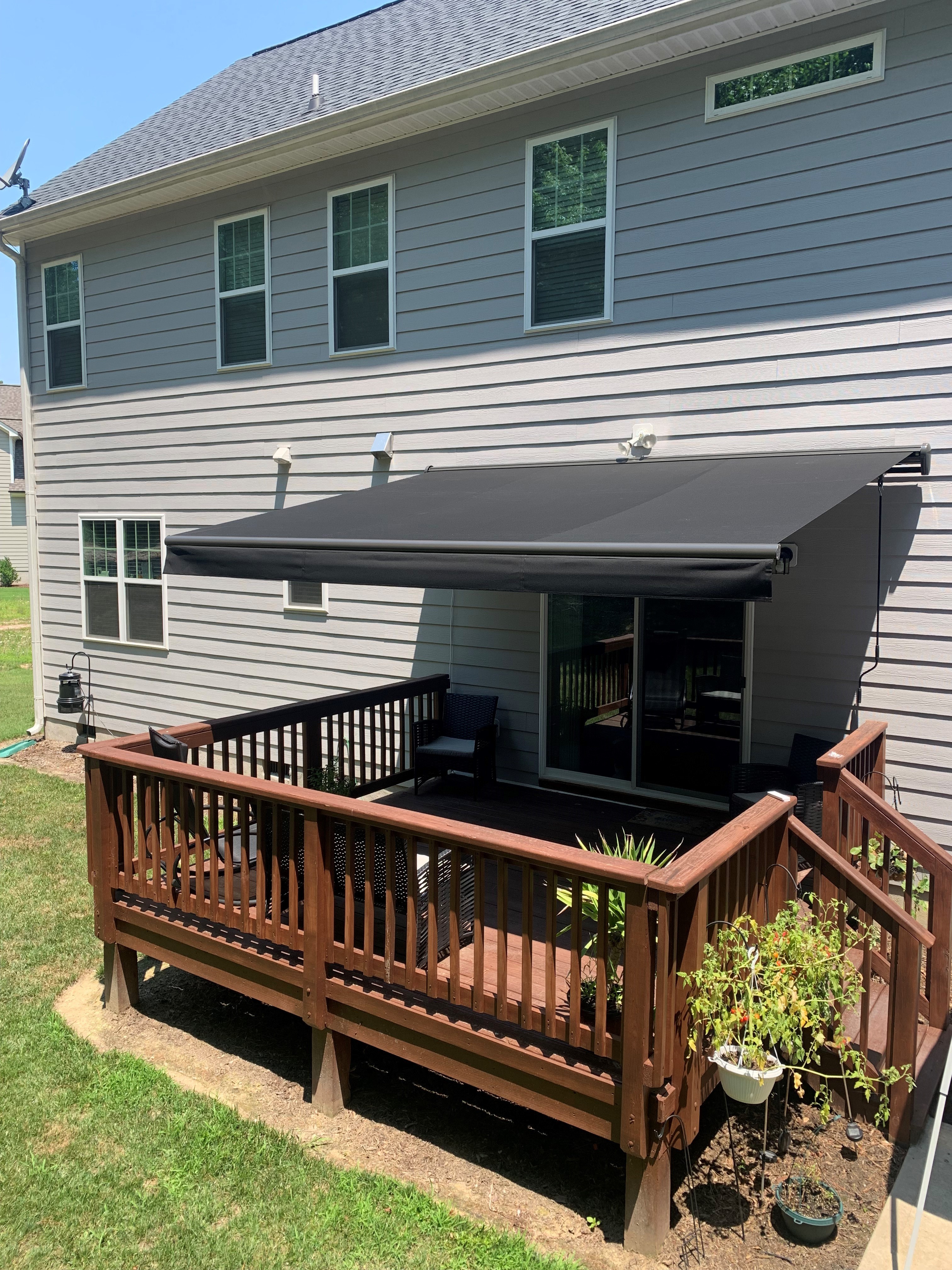 Charcoal gray motorized retractable awning fully extended over a raised wooden deck with black railing on a suburban two-story home