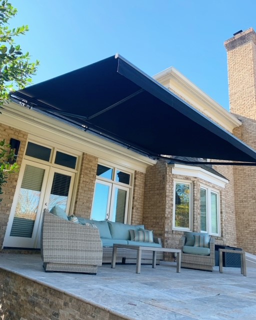 Large navy blue retractable awning fully extended over an elegant stone patio with wicker outdoor sofa set on a brick luxury home