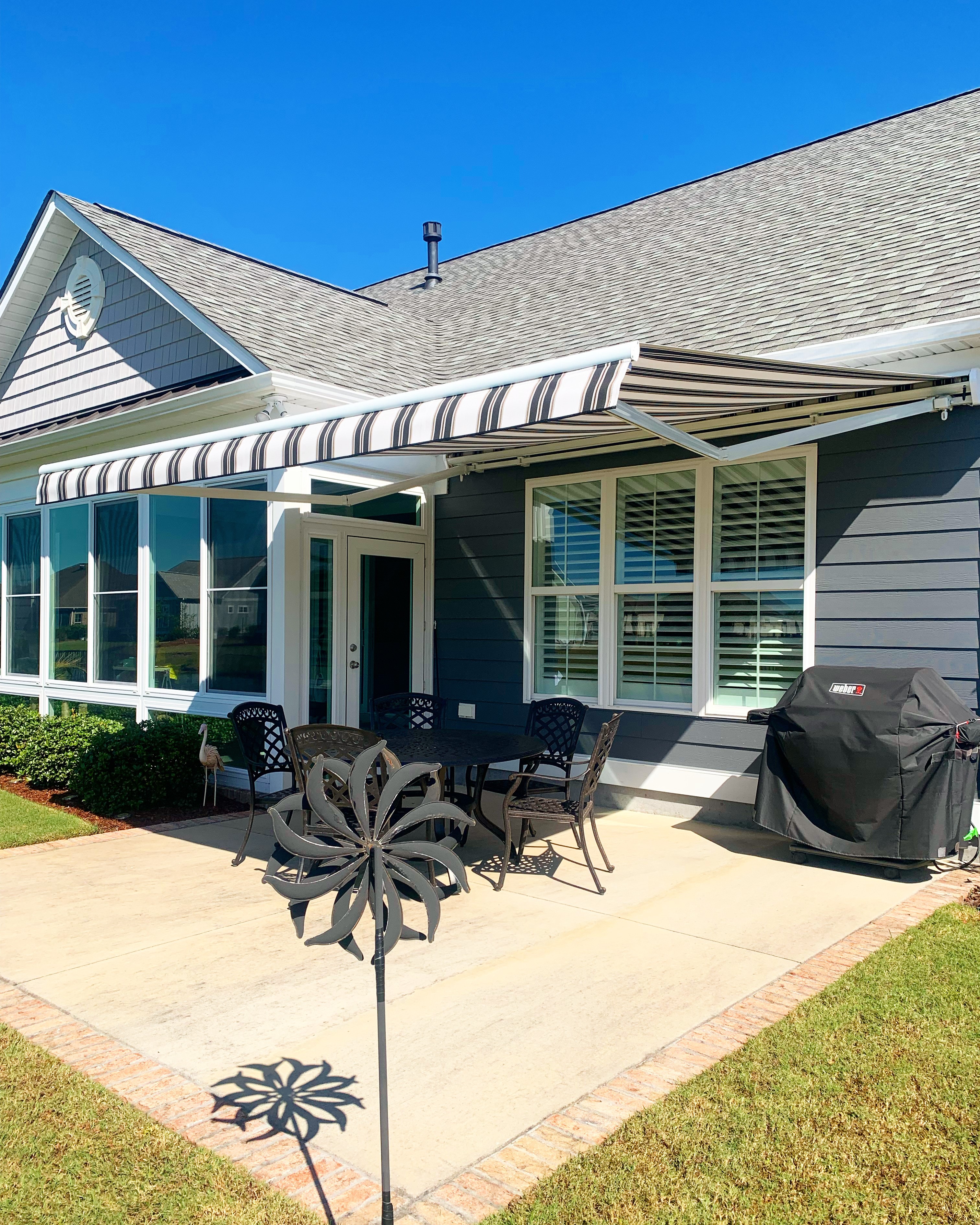 Brown and white striped retractable awning covering a concrete patio with black iron dining table set and outdoor grill on a sunny day
